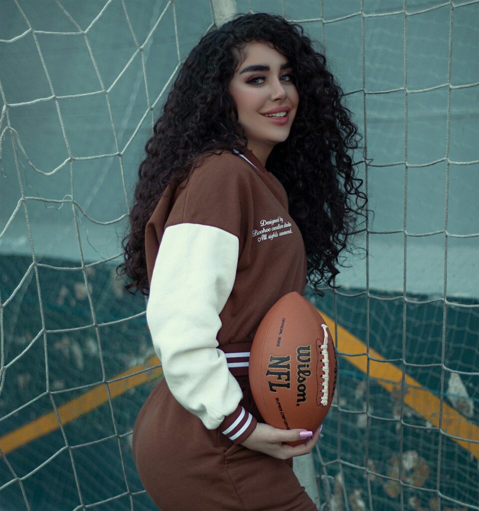 Stylish woman with curly hair smiling and holding a football in front of a net outdoors.